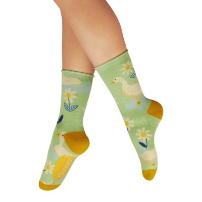 A close-up of a woman's legs wearing a pair of light-green ankle socks with a design of white geese, daisies and puddles, with contrasting ochre heel and toe areas. 