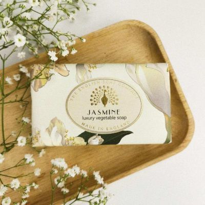 A wrapped bar of soap on a wooden tray that reads, "Jasmine. Made in England." with sprigs of white flowers next to it. 