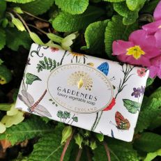 A soap bar wrapped in an illustrated floral wrapper. The label says, "Gardeners luxury vegetable soap." Behind the soap bar are some real pink flowers and green leaves. 