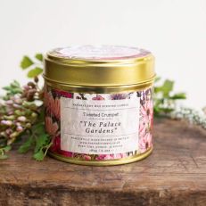 A candle in a gold tin with a lid. The label is illustrate with flowers in pink and purple and the reads, The Palace Gardens. 