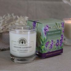A white wax candle in a clear glass container with a label in gold foil that reads English Lavender soy wax candle. Behind is a green and purple box with an illustration of purple lavender and bees. 