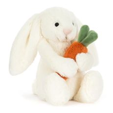 A white soft toy in the shape of a bunny holding an orange carrot with green leaves. With long floppy ears and hard black eyes. 